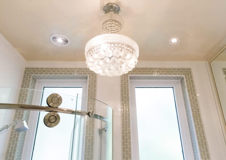 white luxe chandelier in bright white bathroom with green tile surrounding two rectangular windows and top of glass curved shower