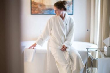 woman in white robe sitting on edge of soaking tub in white marble bathroom at Stonehurst Place