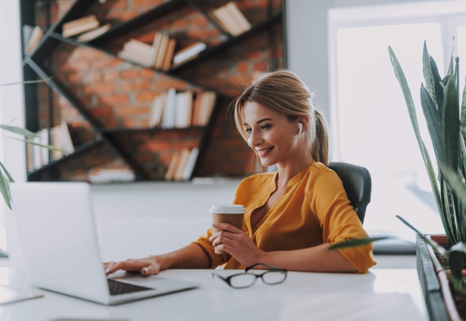 white woman in yellow shirt smiling at laptop holding coffee
